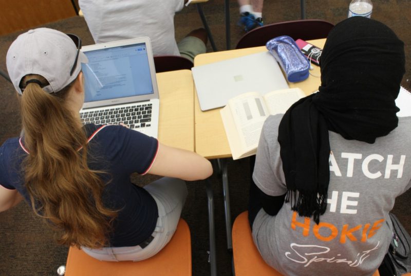 Two girls sit at their desks during class, one wears a baseball cap and the other a hijab.